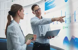 caucasian male and female data scientists with laptop and tablet standing next to big digital screen with graphs and charts in monitoring office. colleagues discussing business opportunities or risks.